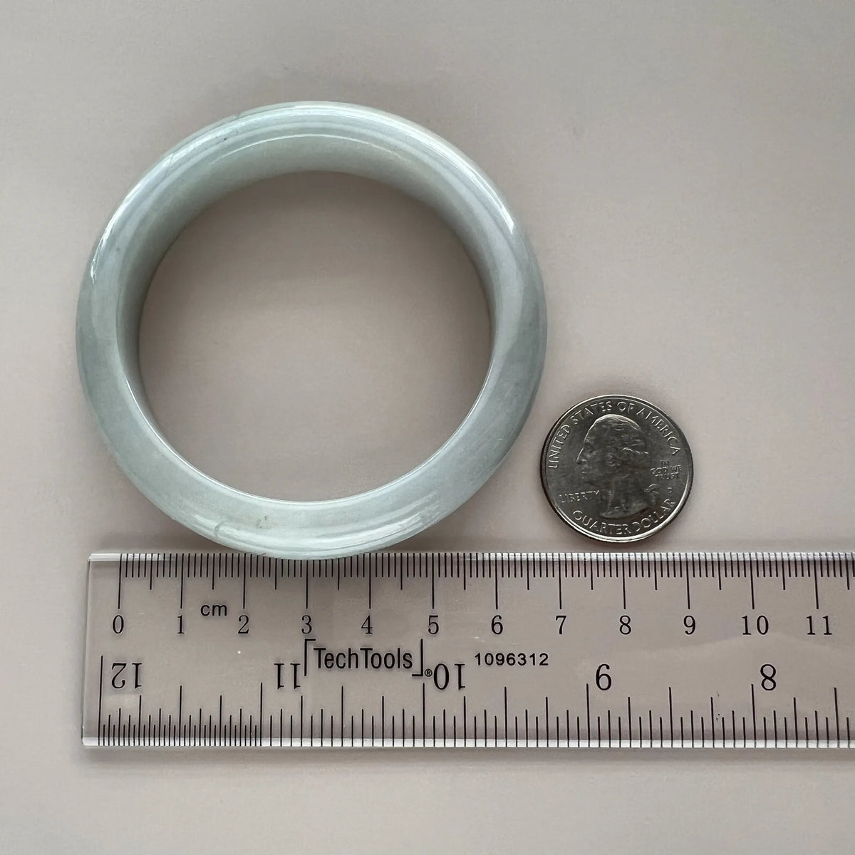 Top-down view of icy light gray jadeite bangle with smooth polished finish beside coin and ruler for scale