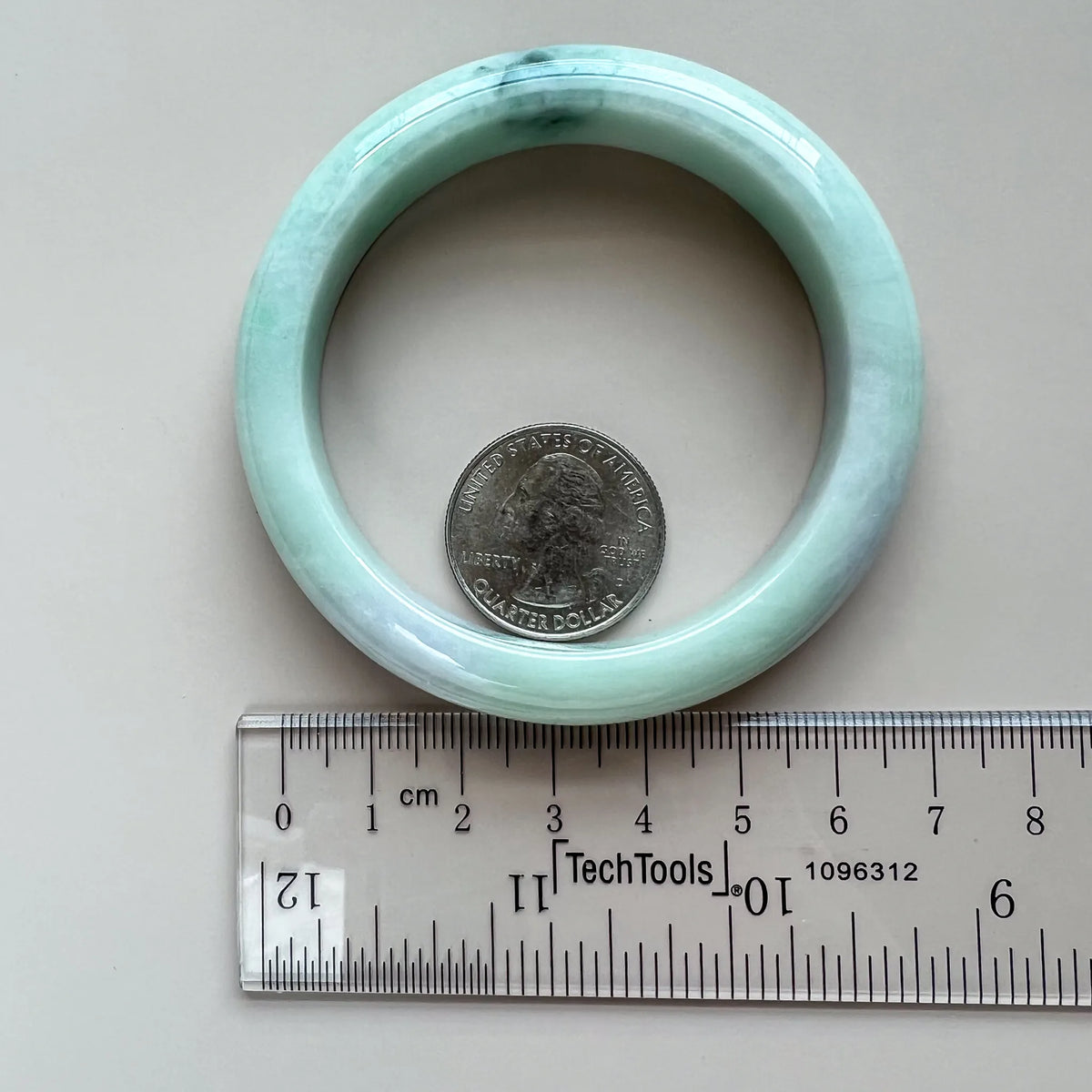 Light green-blue jadeite bangle shown flat beside a ruler and quarter for size reference