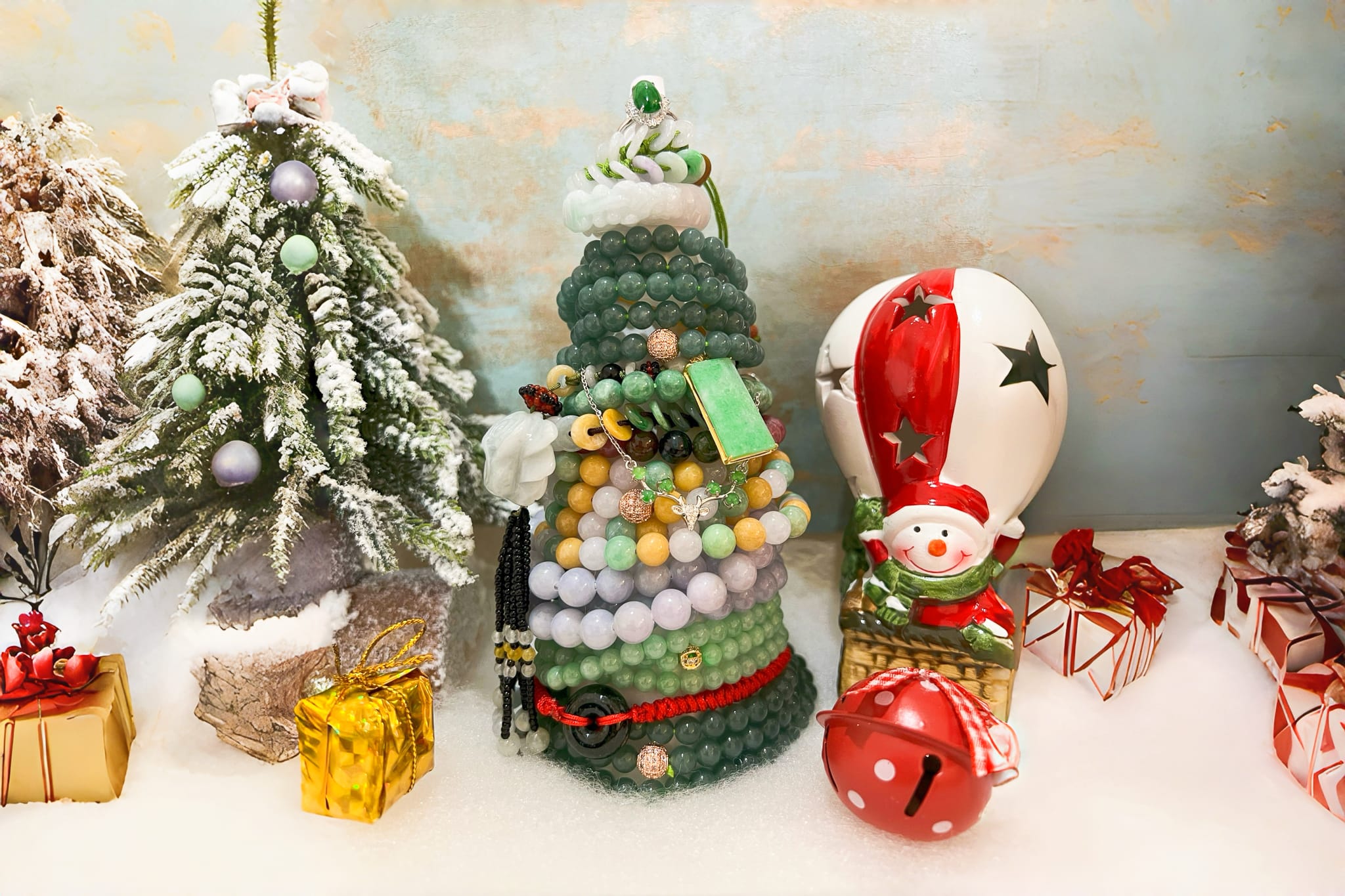 Decorative Christmas tree sculpture created from stacked jade and gemstone jewelry, featuring polished green, white, and lavender jade beads arranged in a layered tree form. The design includes jade bangles, carved jade charms, a rectangular green jade pendant with gold trim, tassel accents, and rose-gold spacer beads, topped with a jade ring. Displayed on faux snow with surrounding holiday décor, showcasing artisan jade craftsmanship and natural stone textures.