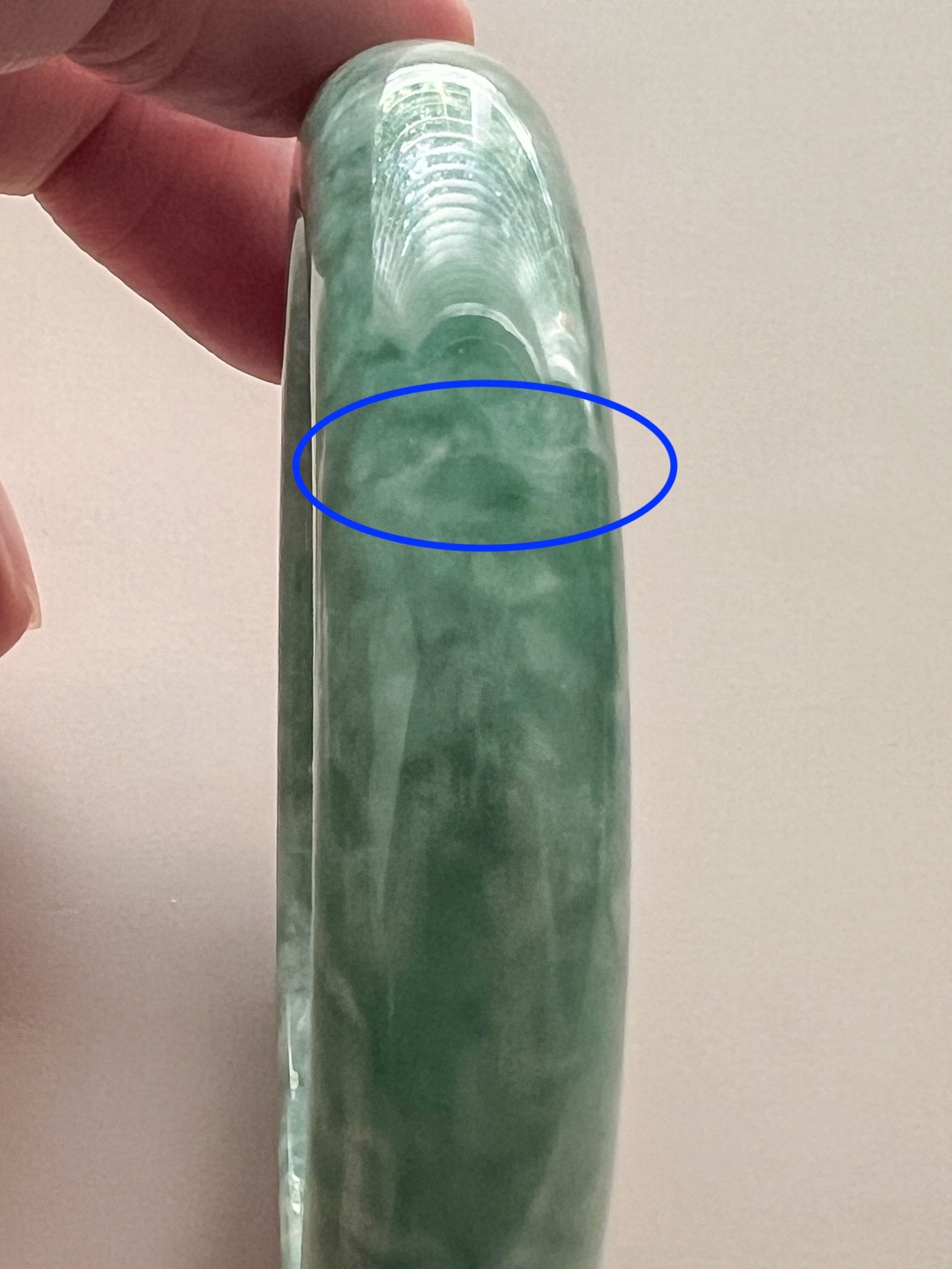 Side view of mottled green jadeite bangle with glossy polished finish and visible cloudy inclusion area highlighted in blue