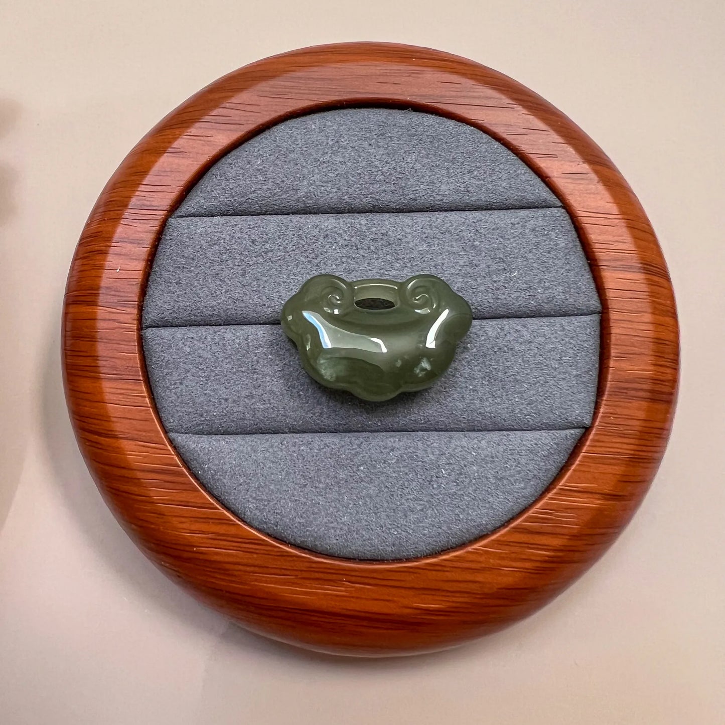 Green nephrite jade ruyi pendant on gray velvet in a round wood display case, glossy carved finish, top view