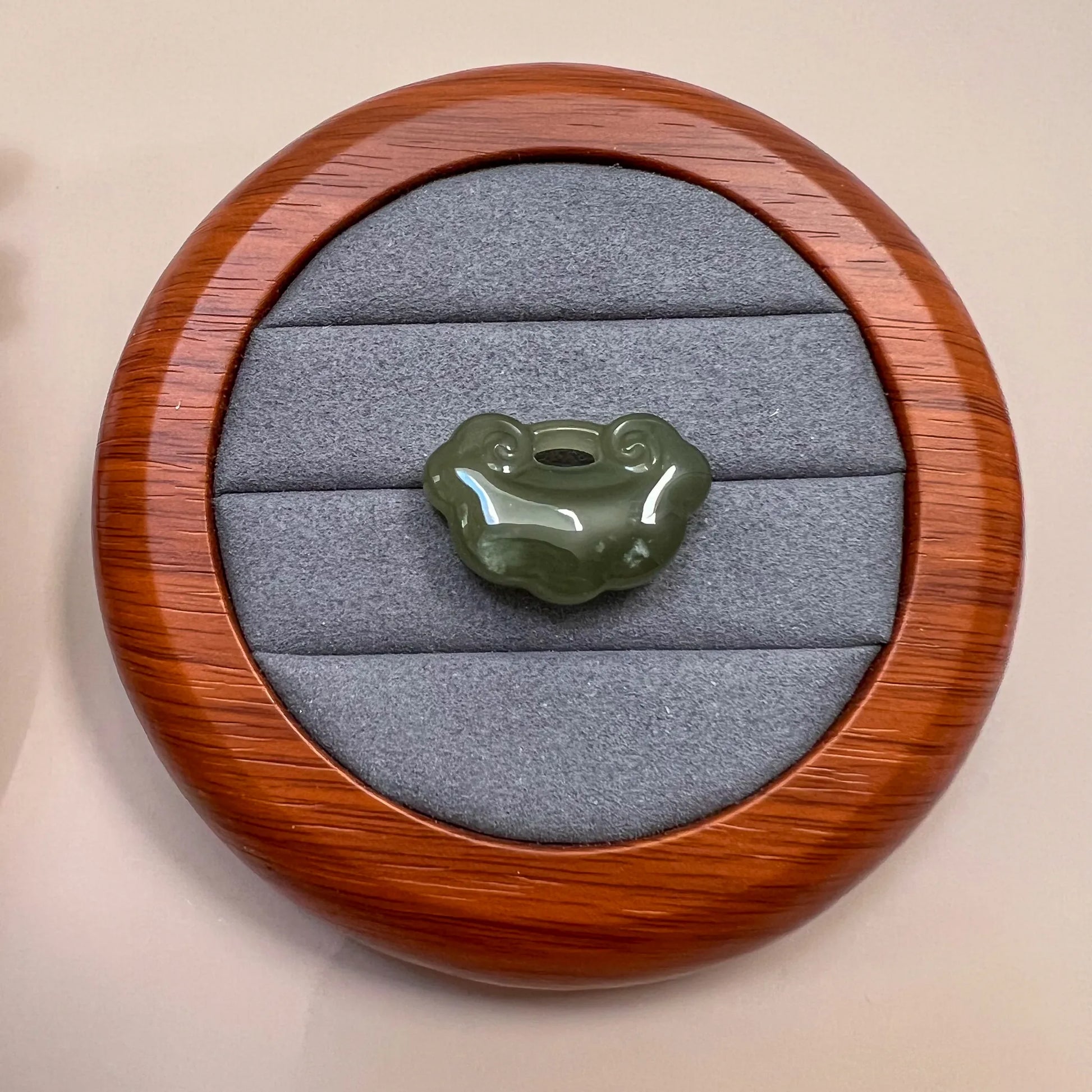 Green nephrite jade ruyi pendant on gray velvet in a round wood display case, glossy carved finish, top view