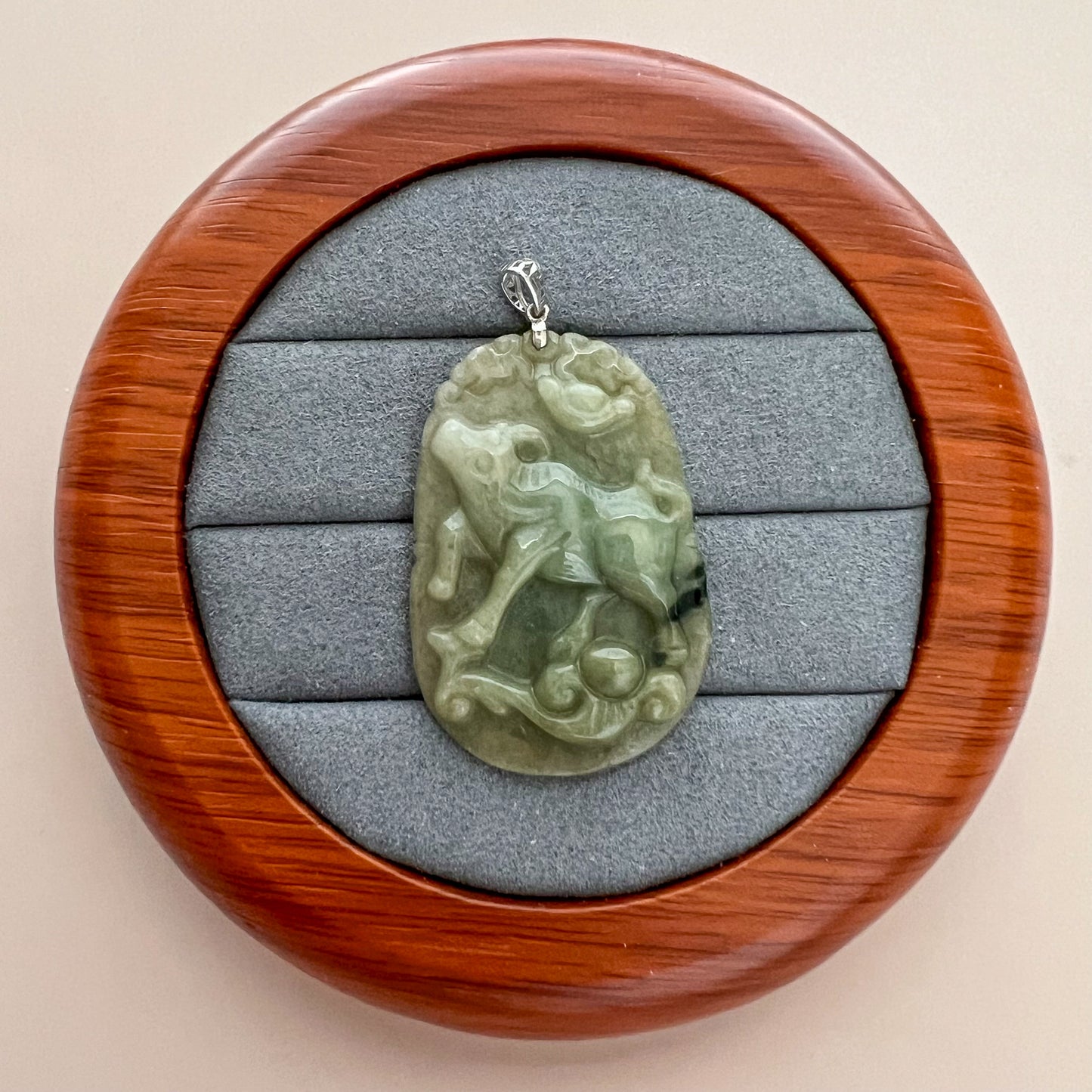 Green jadeite pendant with carved ox and ruyi motifs, sterling silver bail, displayed on a gray jewelry tray in close-up