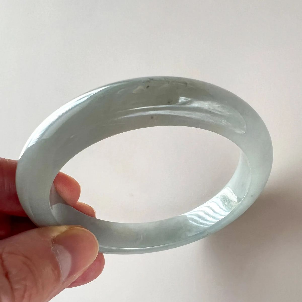 Side-angle close-up of icy light gray jadeite bangle, smooth polished translucent finish, held in hand