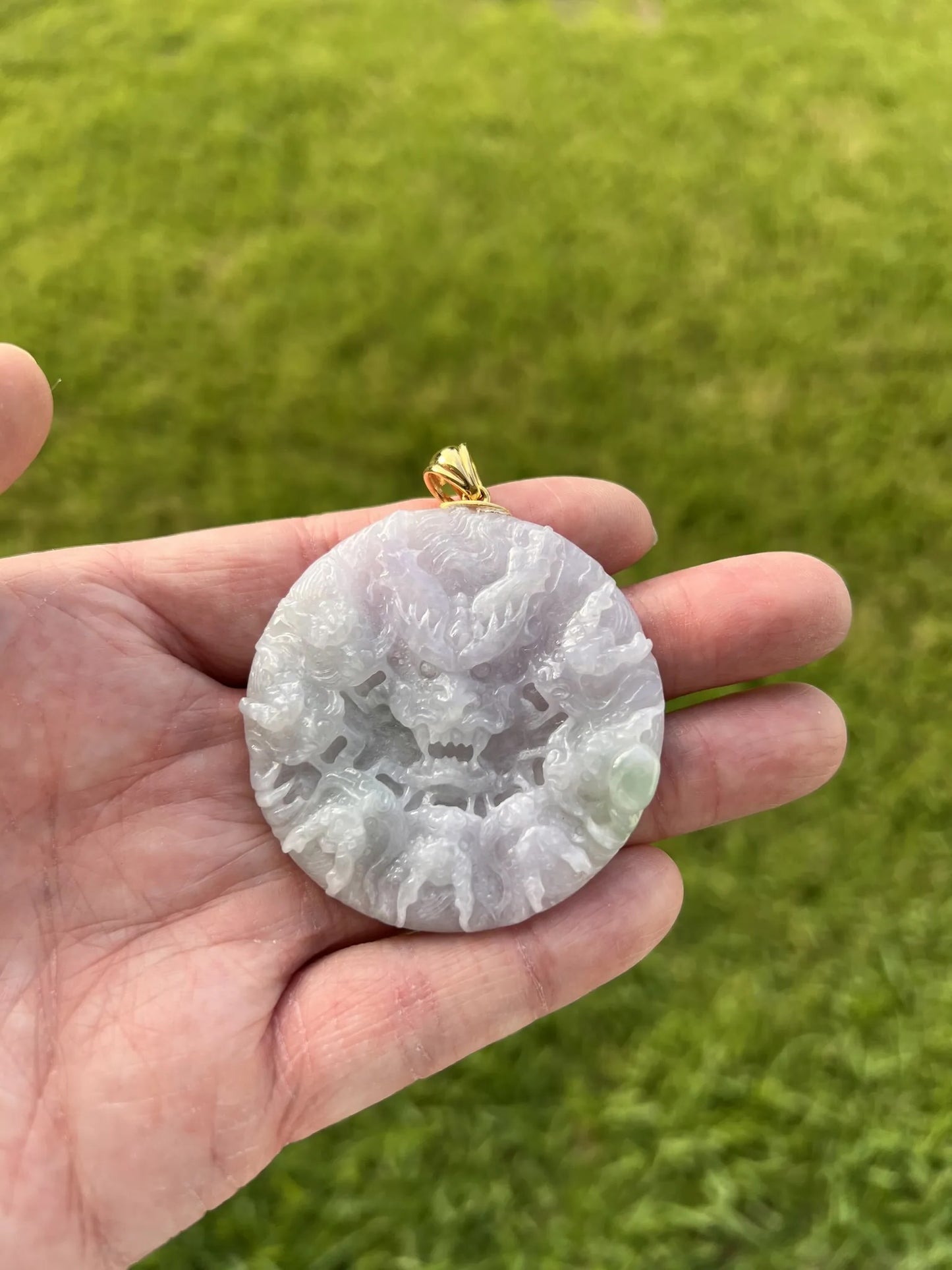 A hand holding a round carved jade pendant with a dragon design, featuring a gold-colored bail, set against a blurred green grass background.