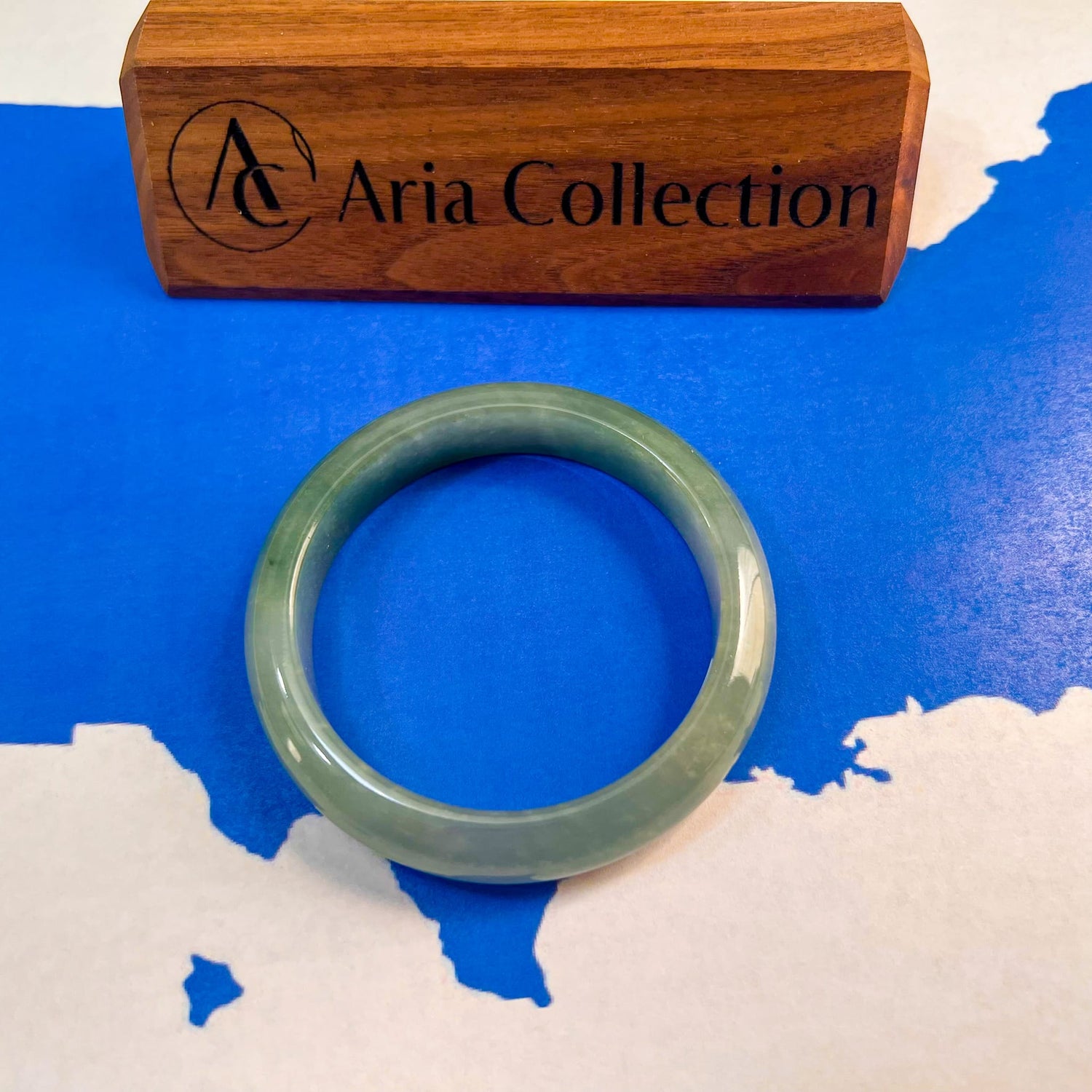 A light green jadeite bangle bracelet displayed on a blue and white map background, with a wooden box engraved with 'Aria Collection' behind it. The background is the map of the USA depicting all products on Aria Design Collection ship from Texas, USA.