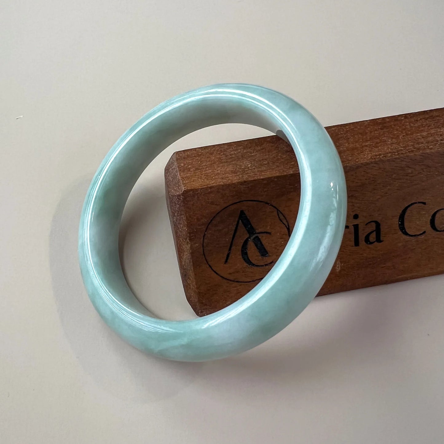 Light green blue jadeite jade bangle angled on a wooden stand, glossy polished finish, smooth classic round profile