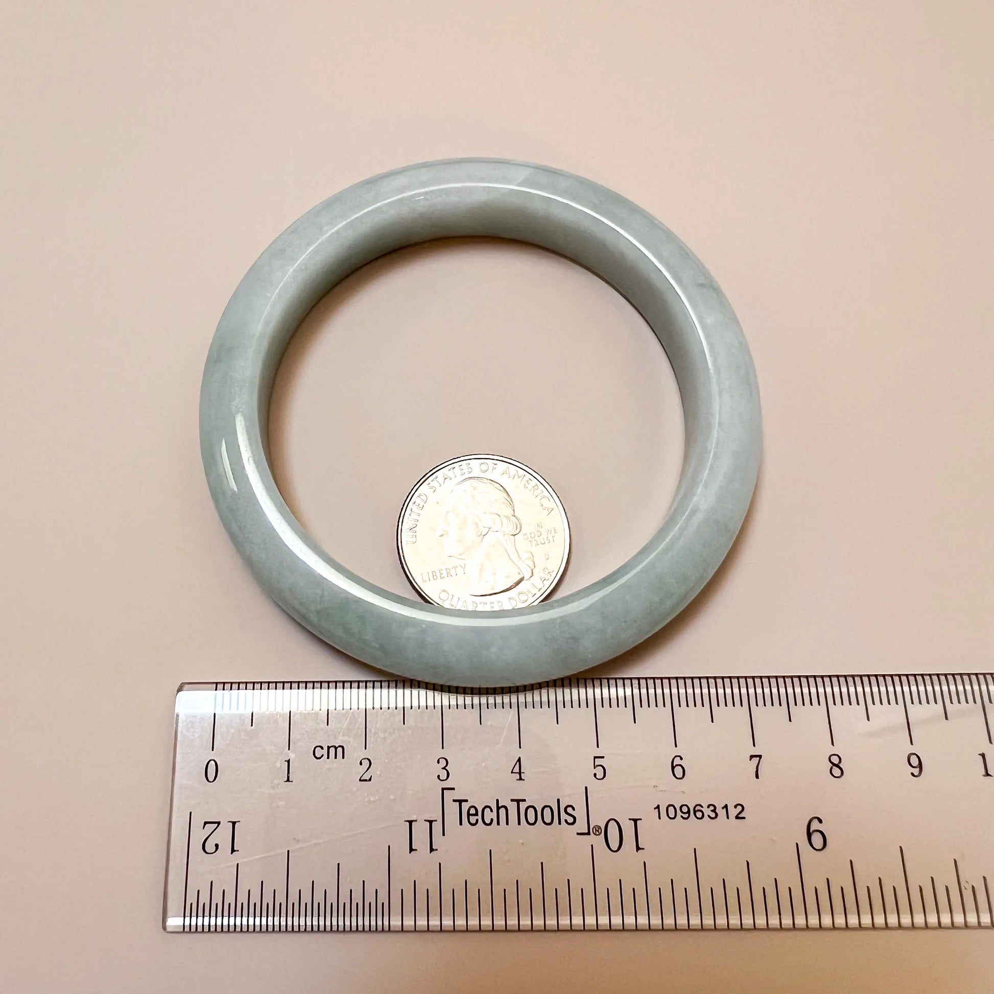 Top-view light green gray jadeite bangle with smooth polished finish beside a quarter and ruler for scale