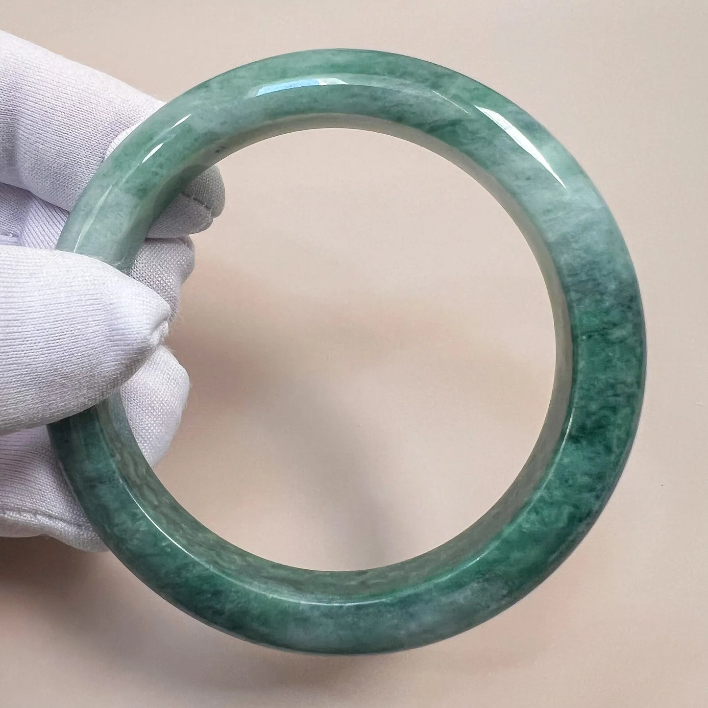 Mottled green jadeite bangle shown at an angle, glossy polished finish, held in white-gloved hand