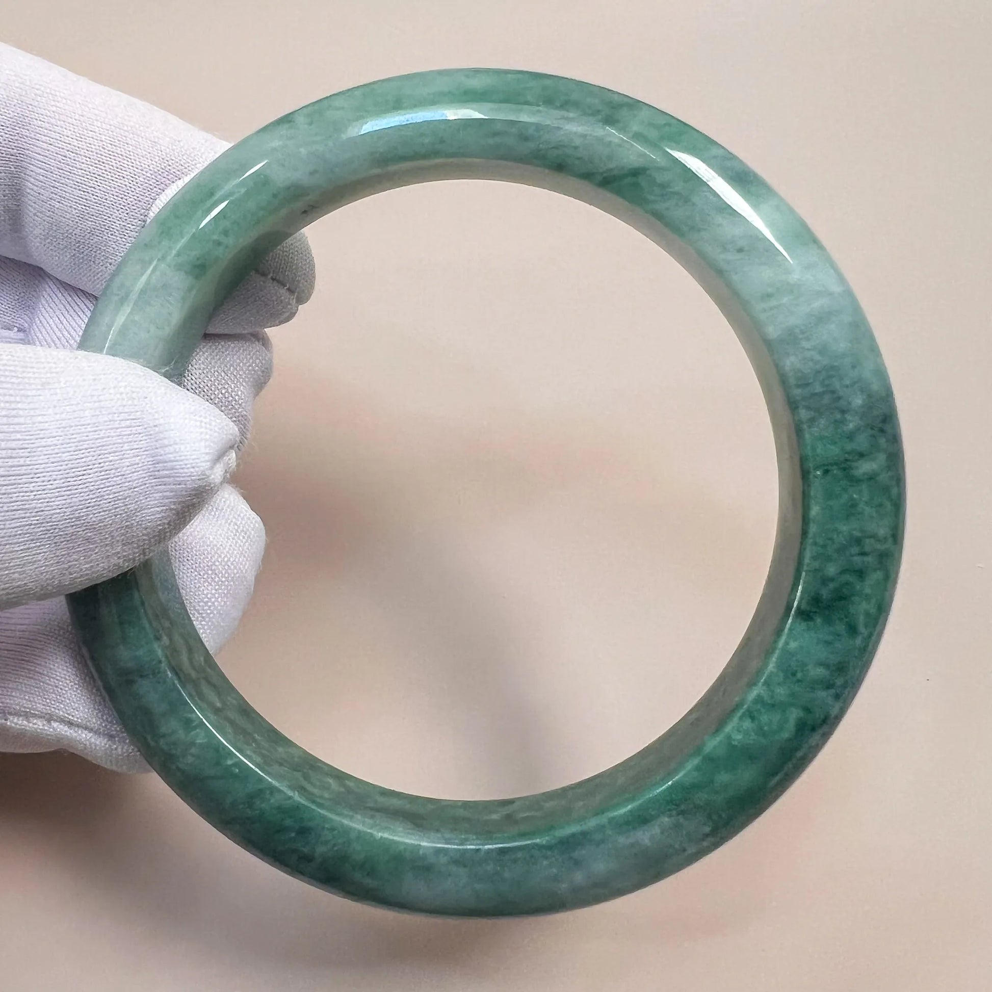 Mottled green jadeite bangle shown at an angle, glossy polished finish, held in white-gloved hand