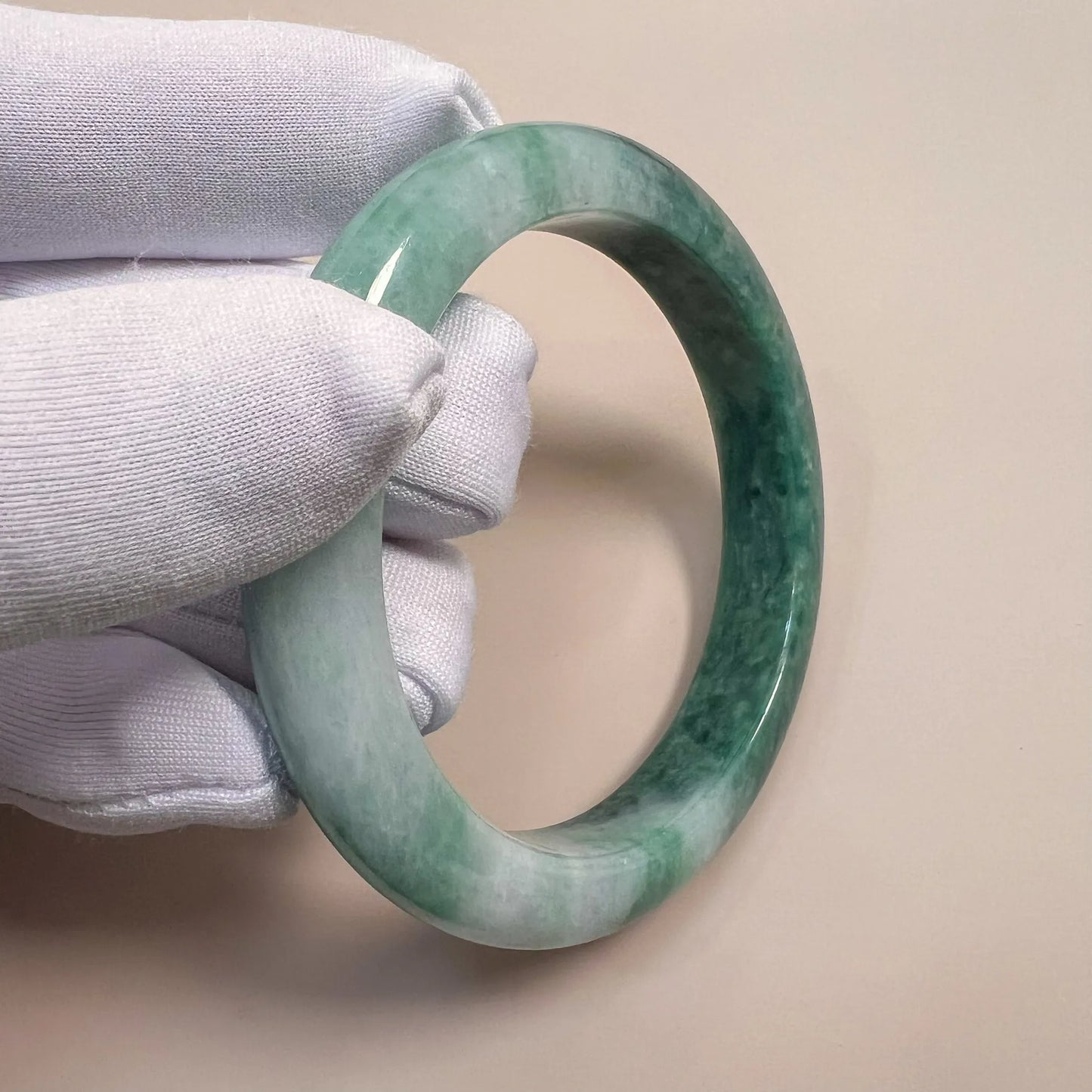 Close-up angled view of mottled green jadeite bangle held in white gloves, showing glossy polished finish and texture