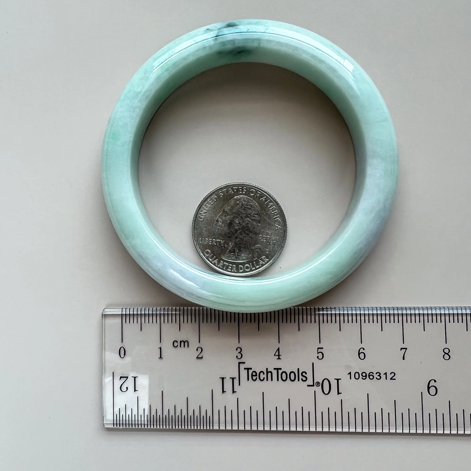 Light green-blue jadeite bangle shown flat beside a ruler and quarter for size reference