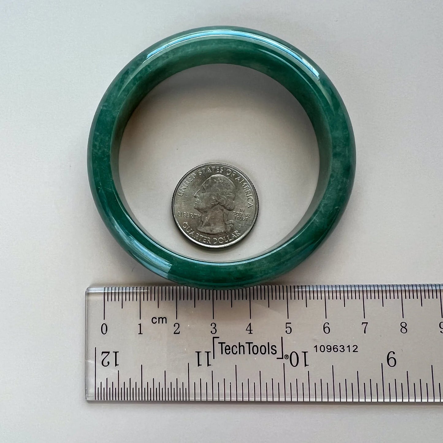 Dark green jadeite bangle shown flat beside a ruler and quarter for scale