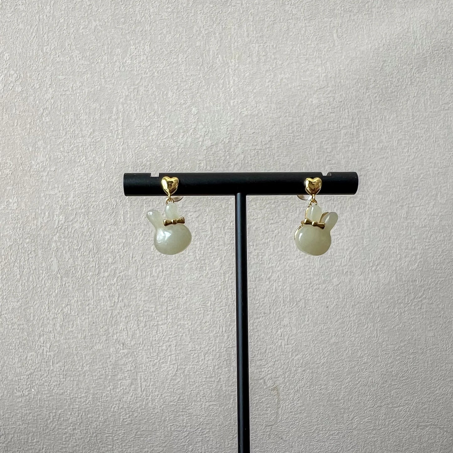 Bunny rabbit nephrite jade stud earrings with gold-plated silver bows on a black display stand against textured wall