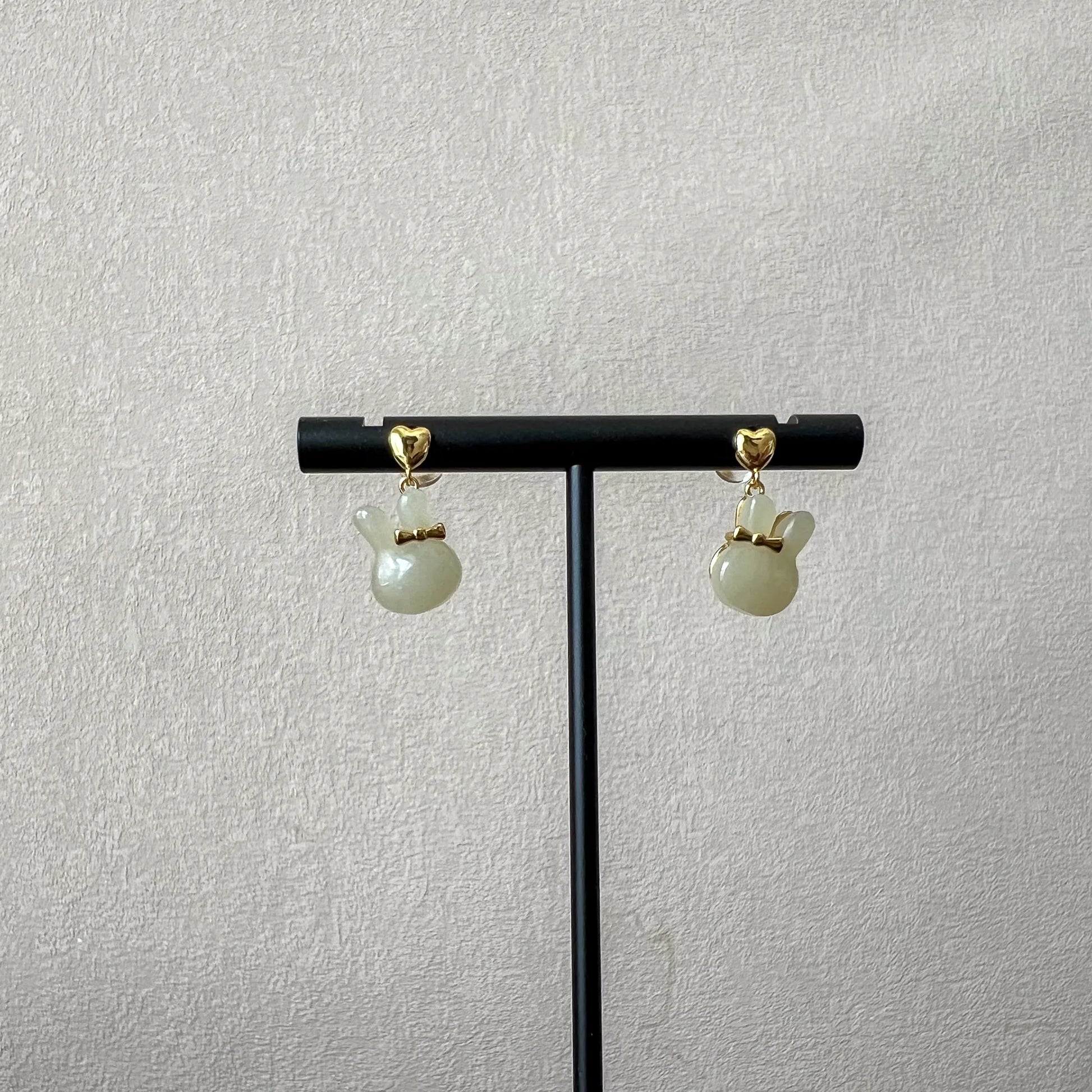 Bunny rabbit nephrite jade stud earrings with gold-plated silver bows on a black display stand against textured wall