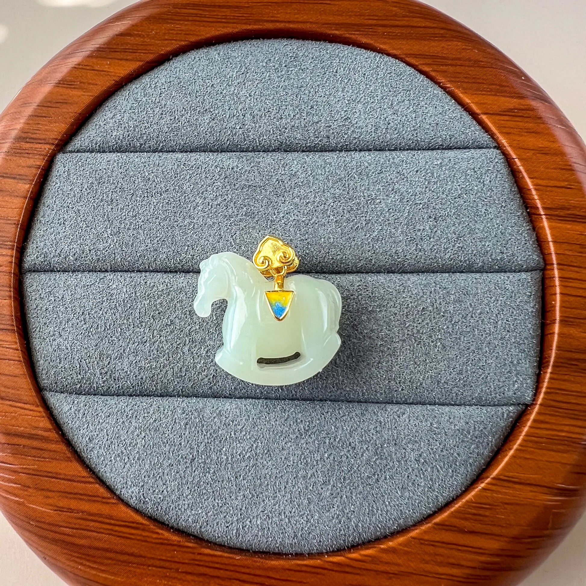 Front view of jade horse pendant in round velvet jewelry box, polished pale green stone and gold detail