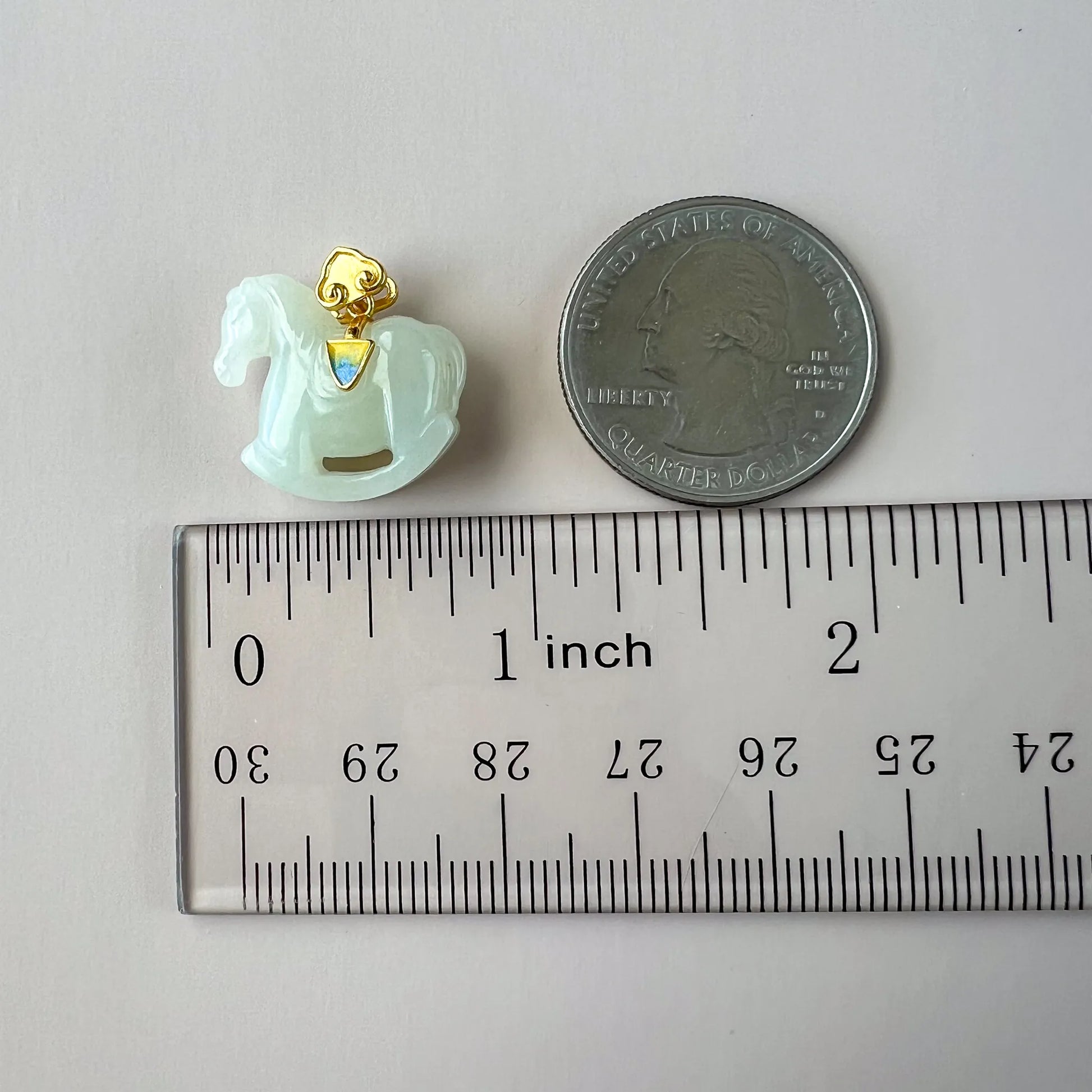 Close view of small light green rocking horse pendant with gold-tone bail beside ruler and quarter