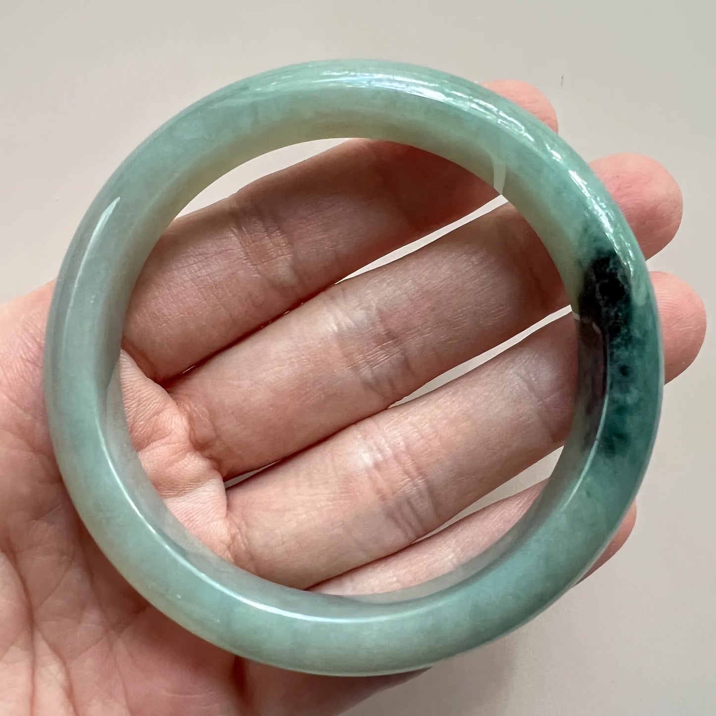 Hand-held view of the rounded jadeite bangle showing translucency, smooth inner surface, and dark green variation