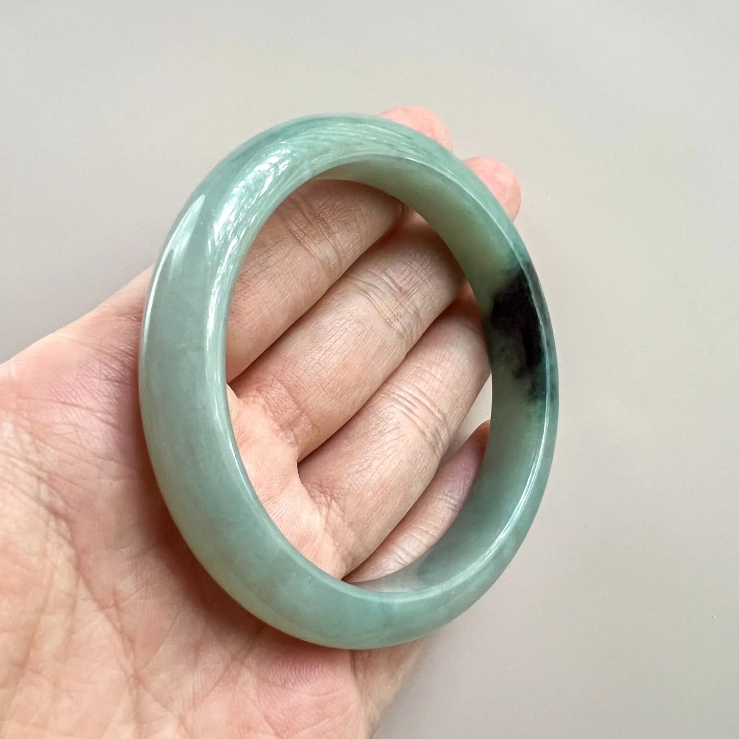 Handheld angled view of polished jade bangle with dark inclusion and translucent finish