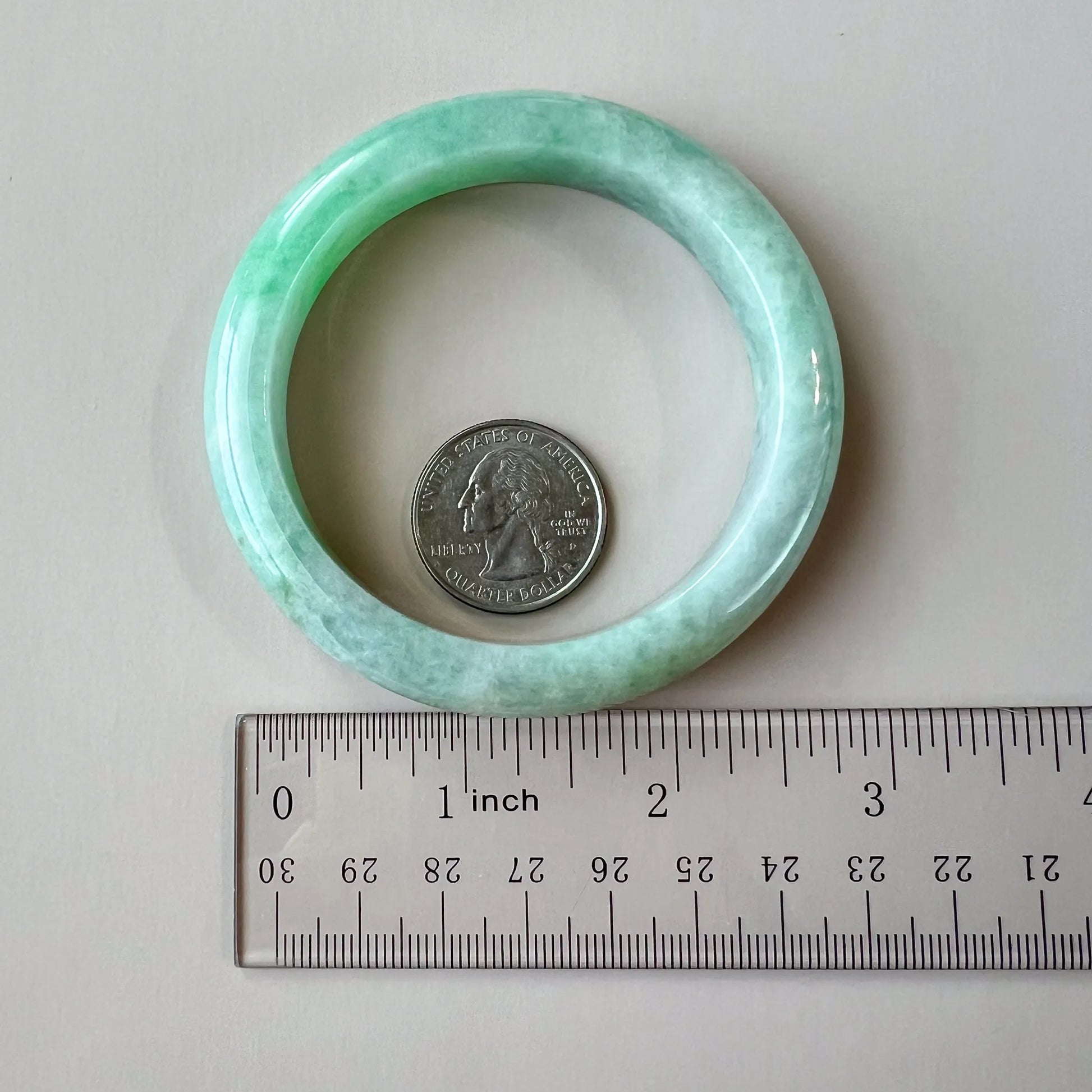 Light green jadeite bangle photographed overhead with quarter and ruler for size reference, polished smooth finish