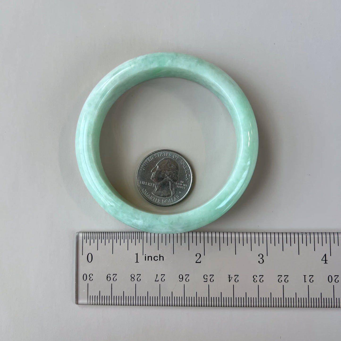 Light green jadeite bangle shown flat beside a ruler and quarter for scale