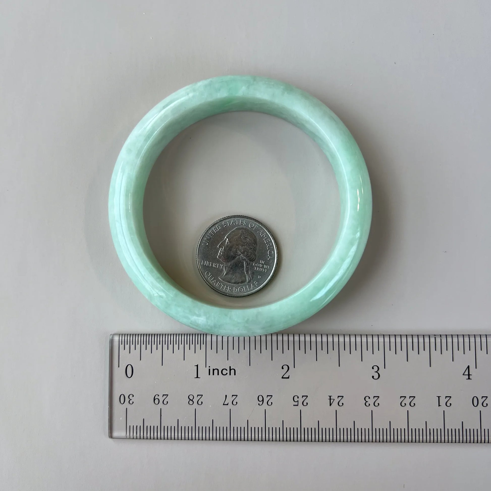 Light green jadeite bangle shown flat beside a ruler and quarter for scale