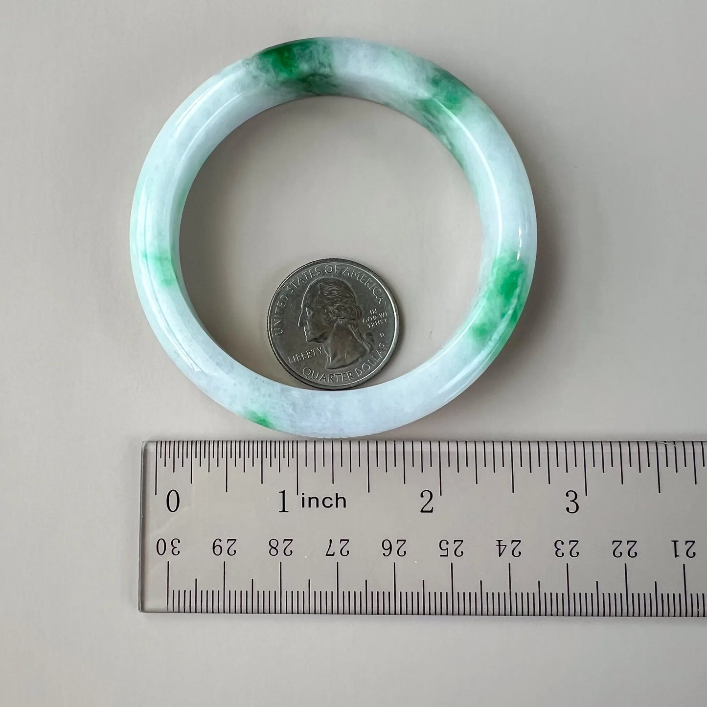 Full view of the icy white and green jadeite bangle beside a ruler and quarter for scale