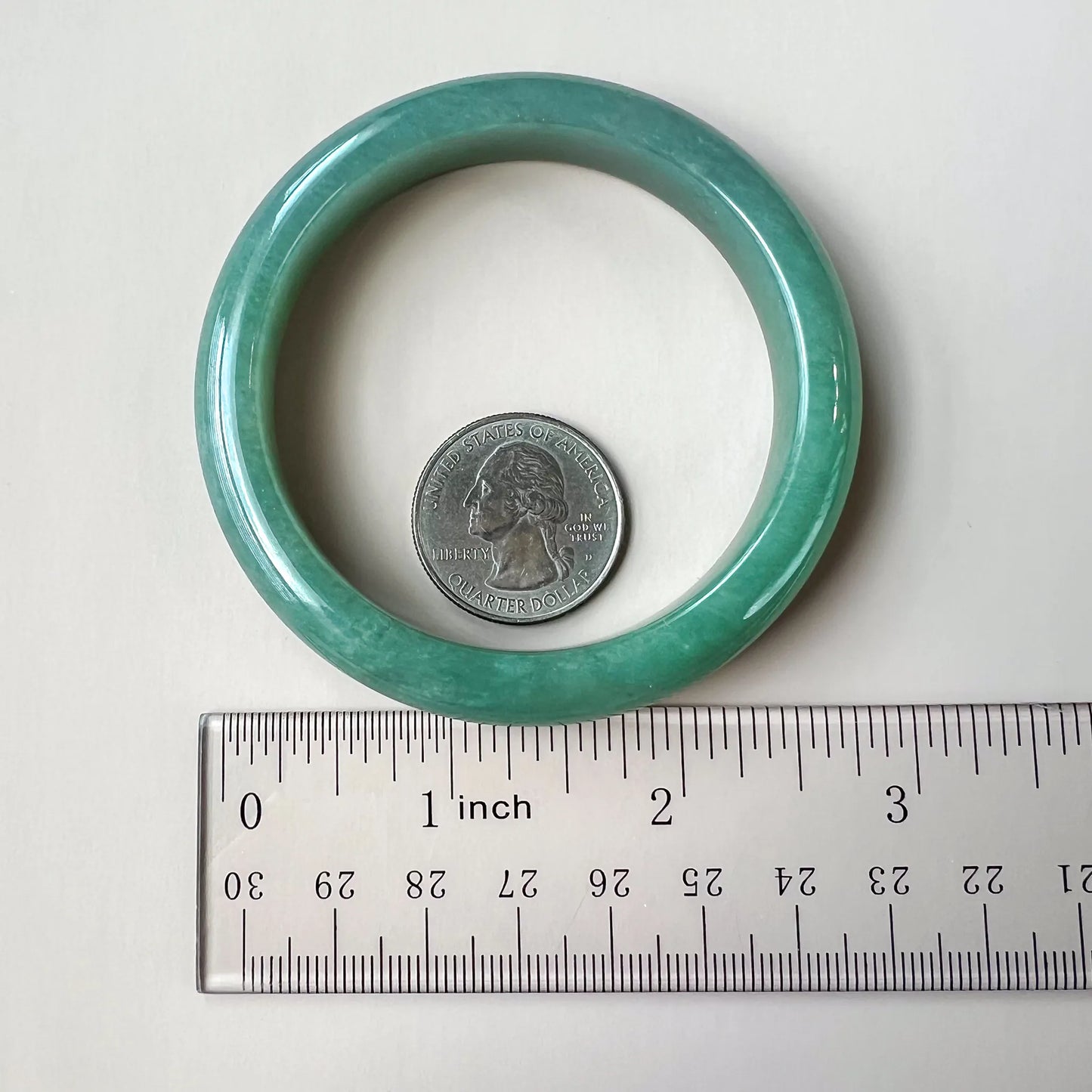 Light green-blue jadeite bangle shown flat beside a ruler and quarter for scale