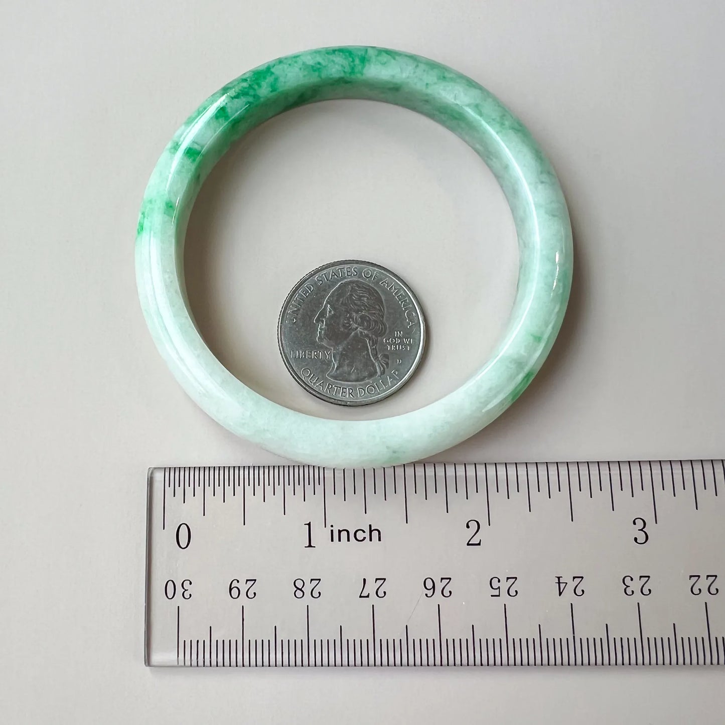 Light green and white jadeite bangle shown flat beside a ruler and quarter for scale