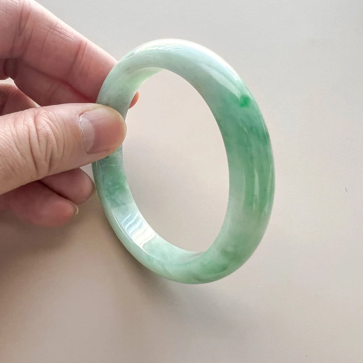 Close-up angled view of a light green jadeite bangle with glossy polished finish and translucent marbled texture in hand
