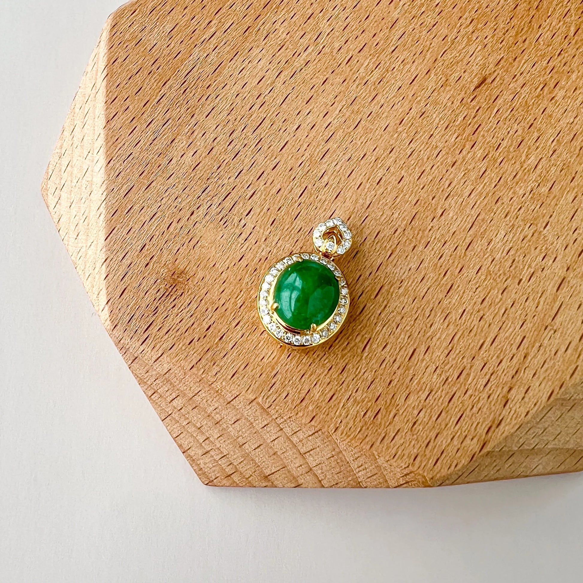 Front view of oval green gemstone pendant on a wood display, gold setting with diamond halo