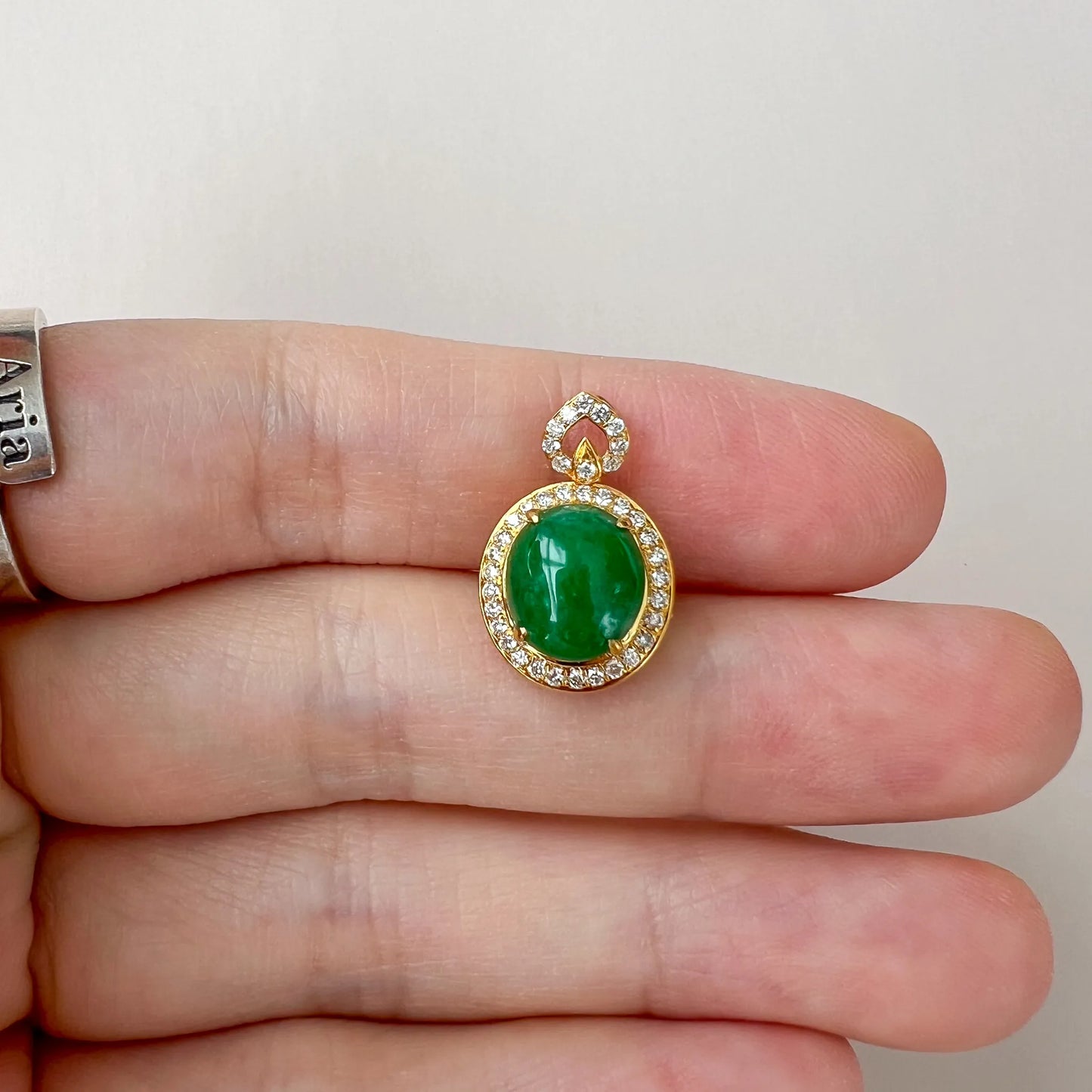 Pendant held on fingers, showing polished green cabochon stone and diamond halo in gold