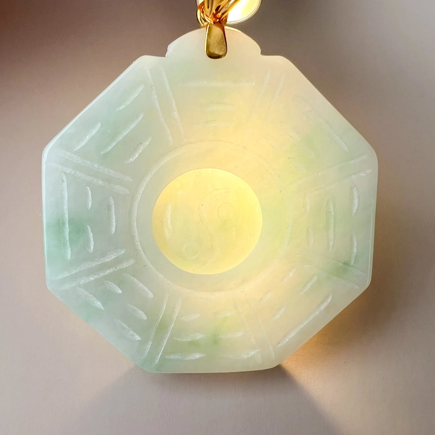 Backlit view showing translucent light green stone, octagonal carving, central yin-yang and gold-tone bail