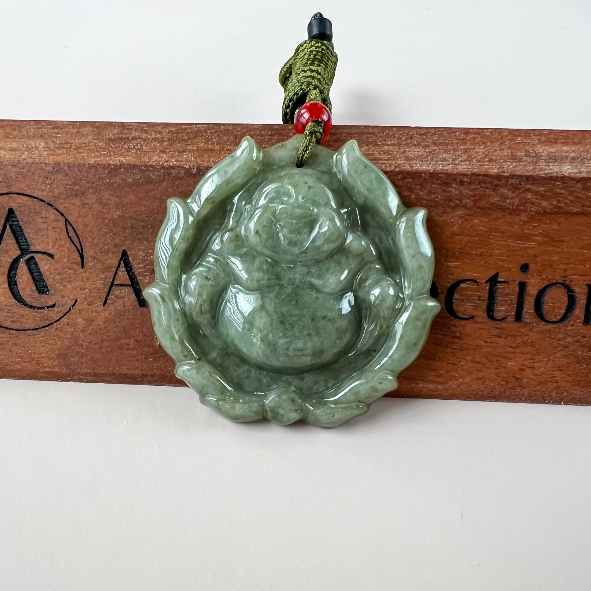Green jadeite jade Happy Buddha lotus pendant, glossy carved front view on cord with red bead against wood background