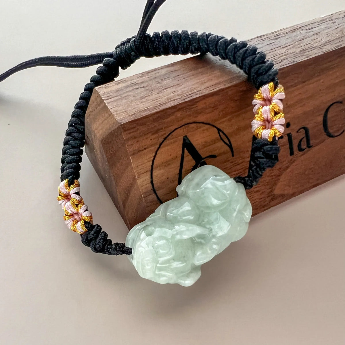 Angled view of carved pale green jade charm on black woven cord with gold accents