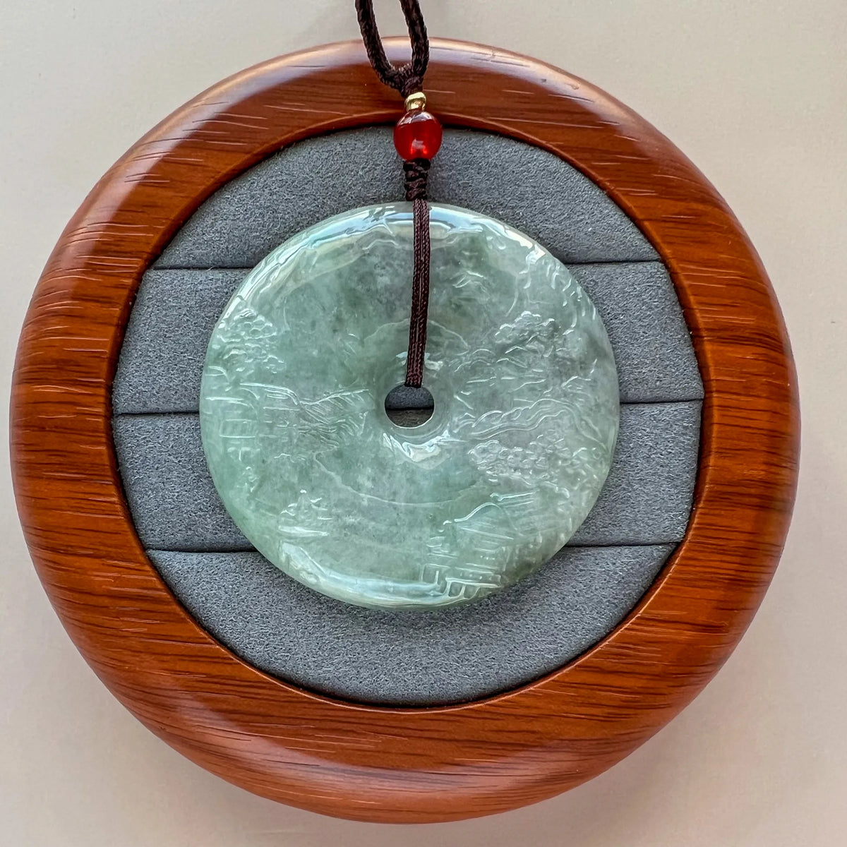 Front pendant displayed on round wood stand with gray padded insert, showing carved jade disc and cord