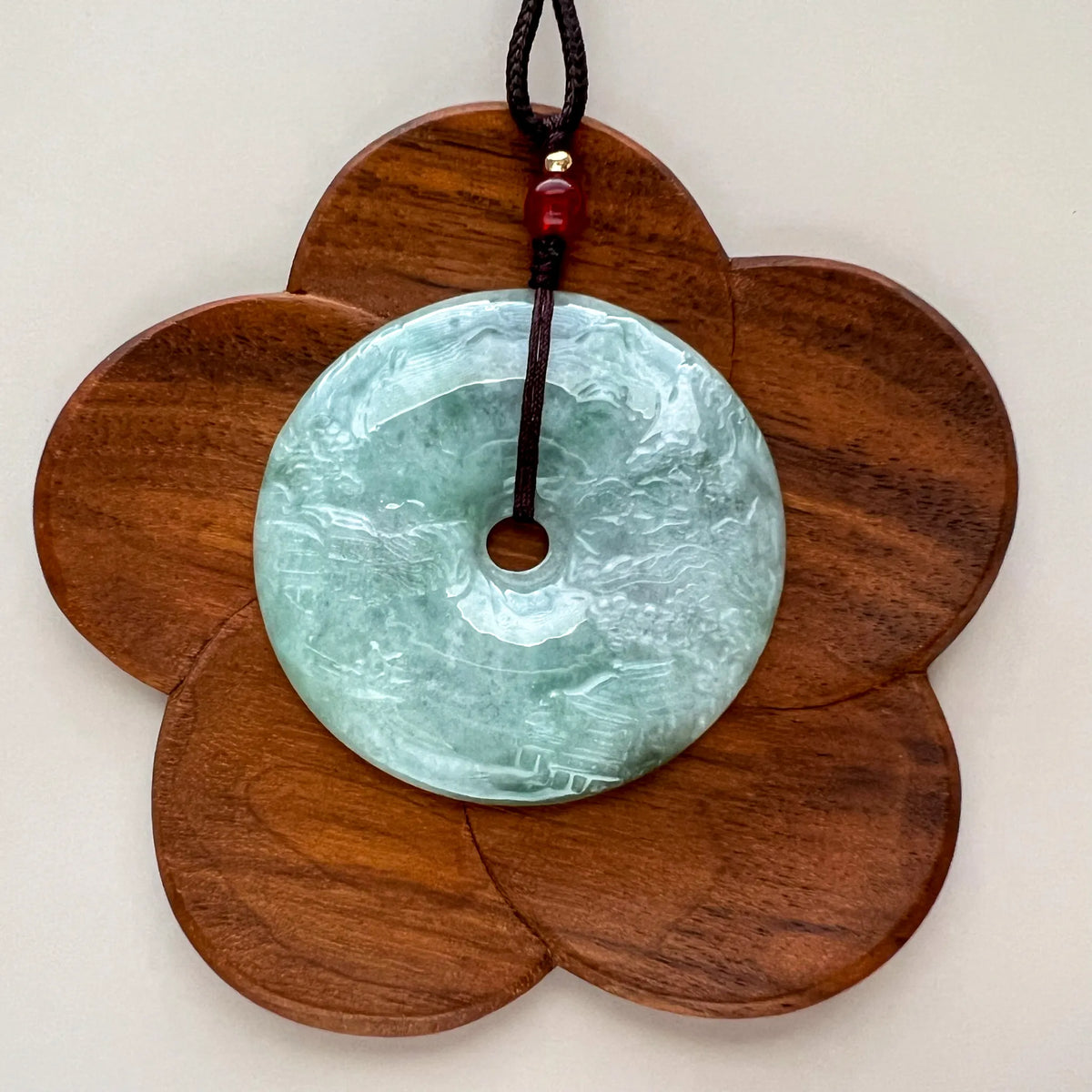 Carved jade disc pendant shown on flower-shaped wood display stand with dark cord and red bead