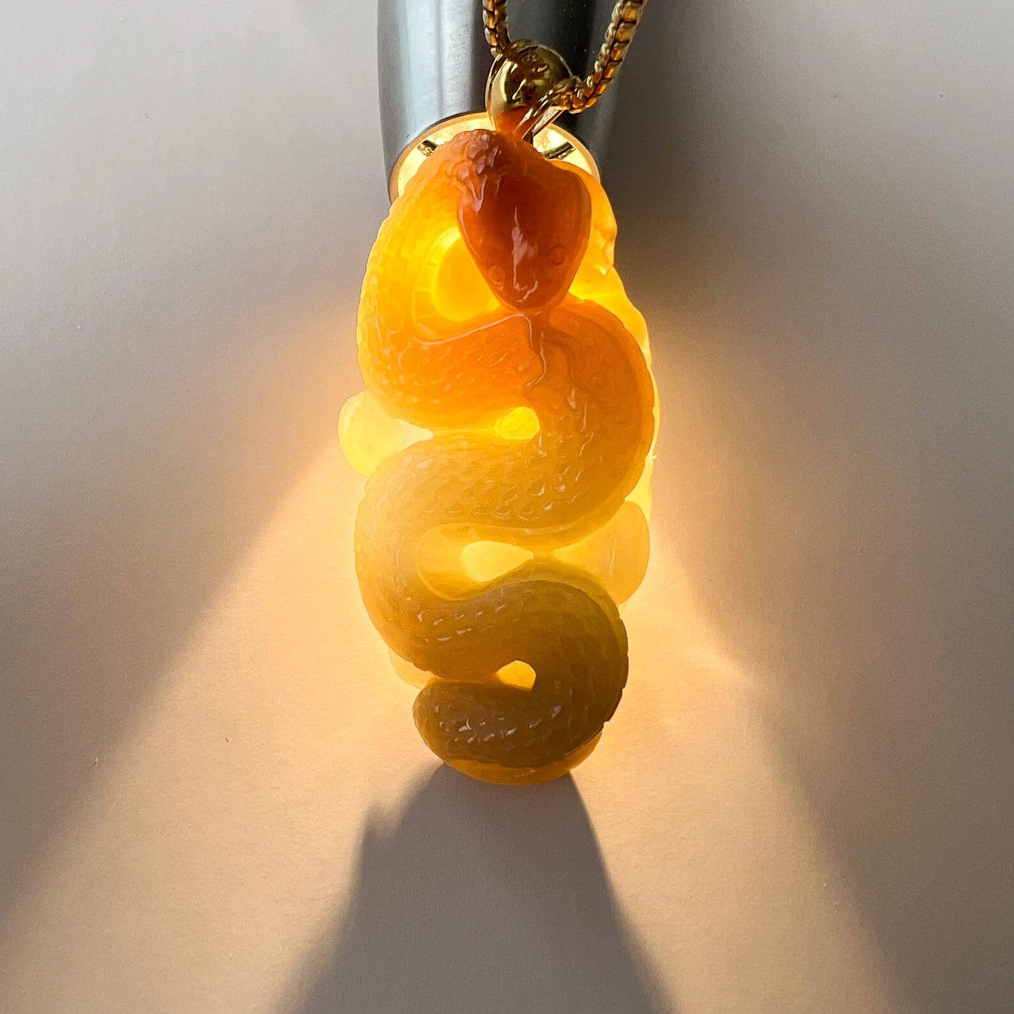 Backlit view of translucent jade snake pendant on chain, glowing amber-green carved finish