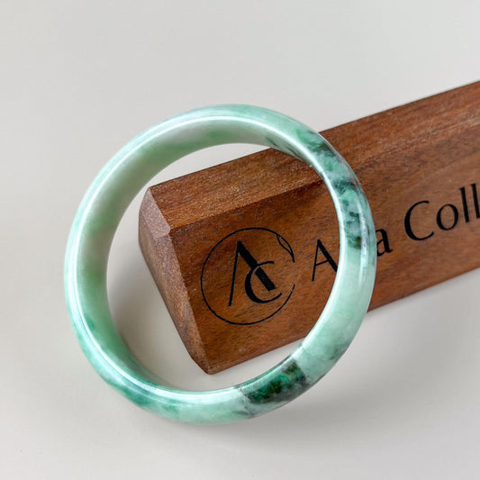 Green Burmese Type A jadeite jade bangle, polished translucent finish, angled close-up on wooden stand with mottled texture