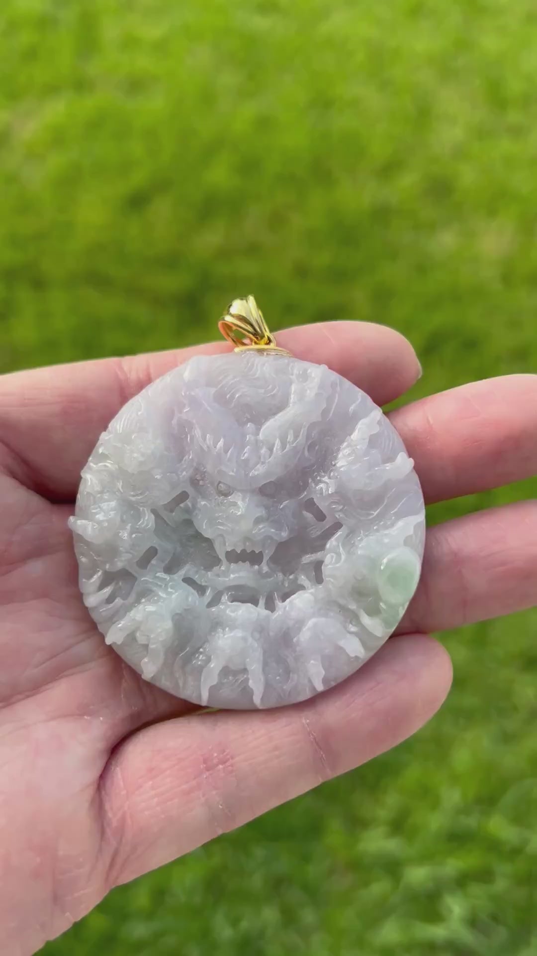 Video of a hand holding a round carved jade pendant with a dragon design, featuring a gold-colored bail, set against a blurred green grass background.