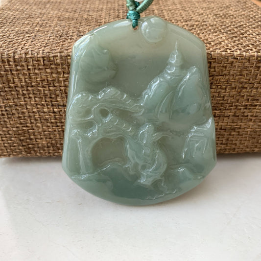 Close-up of pale green jadeite carved landscape pendant with glossy finish and woven cord on textured mat background