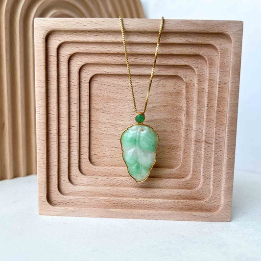 Green jade leaf pendant with solid 18K gold frame on fine chain, displayed on carved wooden stand, close-up front view