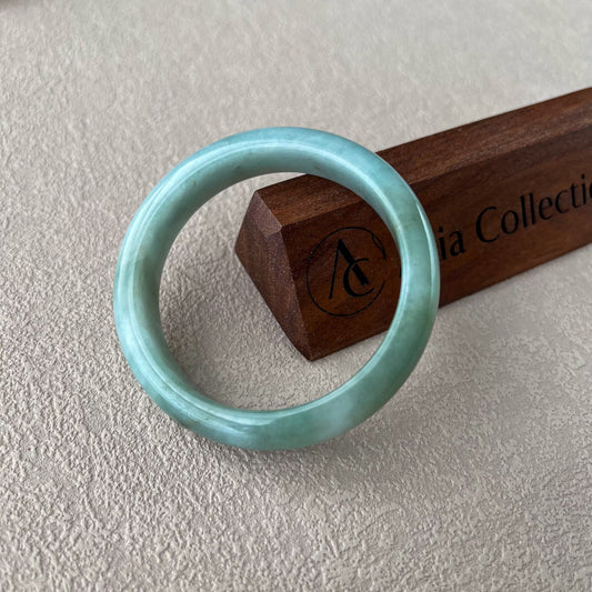 Green Burmese type A jadeite jade bangle shown at an angle, smooth polished finish, on textured fabric with wooden stand